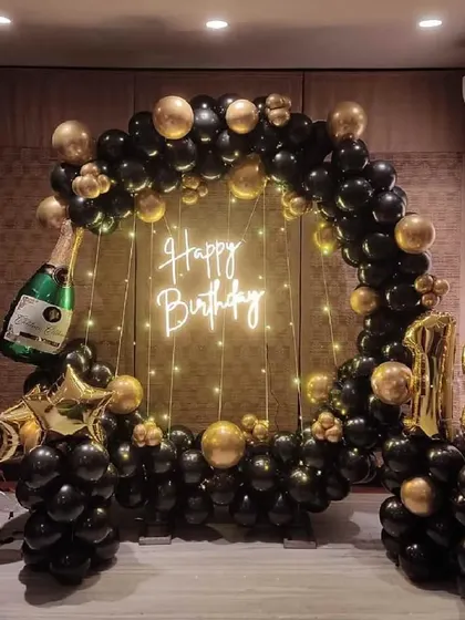 An elegant 18th birthday decoration in black and gold. The circular balloon arch is accented with a champagne bottle prop and a 'Happy Birthday' neon sign for a sophisticated celebration.