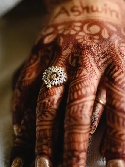 A close up of the bride's hand, showing off her engagement ring and the rich color of her mehendi, with the groom's name written on it.