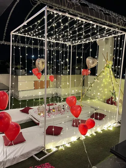 An intimate and romantic cabana setup on a terrace. The space is defined by a frame draped in fairy lights, with low seating, red balloons, and a small tent for a cozy feel.