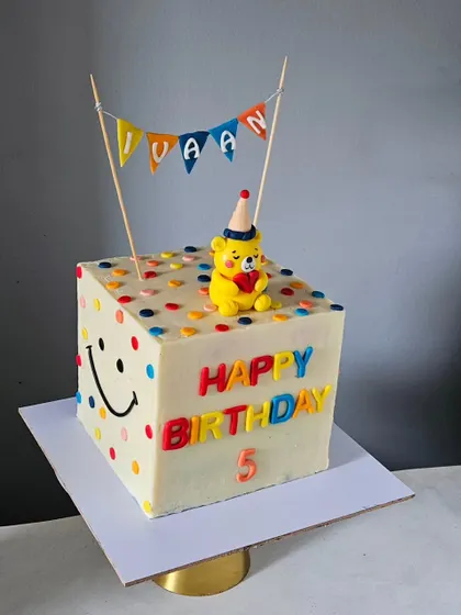A fun and playful cube-shaped cake. This unique design is decorated with colorful confetti dots, a cheerful smiley face, and a cute party bear topper, all on a non-fondant buttercream base.