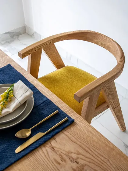 A detail of the dining chair and place setting. The elegant curve of the wooden chair, the pop of yellow from the cushion, and the brass-finished cutlery all contribute to the room's sophisticated yet inviting feel.