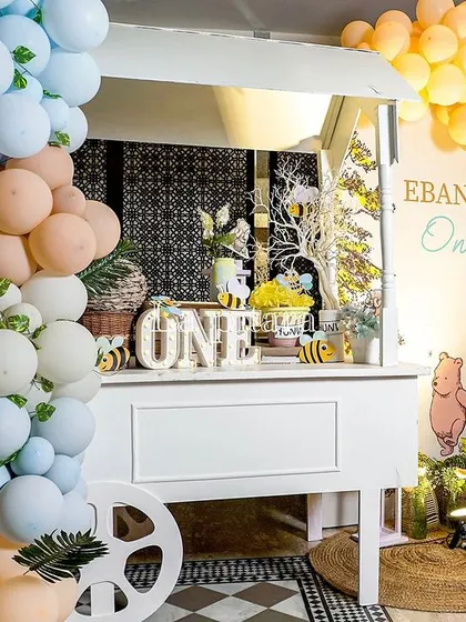 A cute party cart decorated with pastel balloons and a 'ONE' sign, serving as a dessert or favor station for a Pooh-themed first birthday.