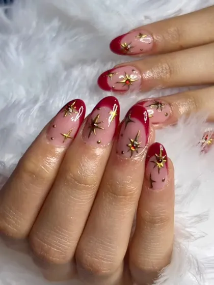 A starry and festive design. This manicure features red French tips on a sheer nude base, adorned with a cascade of gold stars and studs.