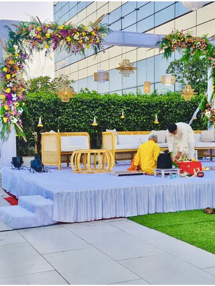 A modern and minimalist vidhi mandap for an outdoor daytime ceremony. The setup features a beautiful floral arch, comfortable cane furniture for the rituals, and elegant hanging lamps.