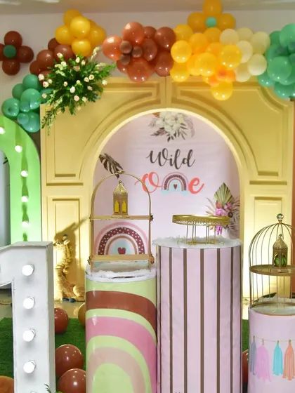 This "Wild One" theme uses a boho-chic color palette. The setup includes a golden arch backdrop, rainbow-printed pedestals, and a variety of animal props for a trendy first birthday.