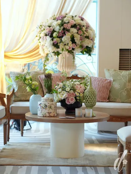 A comfortable lounge area designed with elegant wooden furniture and soft cushions in pastel green and pink. Large floral arrangements add a touch of grandeur to the relaxed setting.