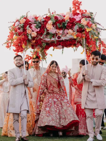 The bride's grand entrance under a lush floral phoolon ki chadar. The vibrant red of the flowers and her lehenga create a stunning visual against the outdoor setting.