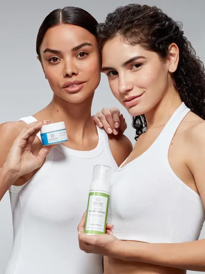 A clean and fresh group shot for a skincare line. Two models in simple white tops hold different products, highlighting the brand's range in a bright, minimalist studio setting.