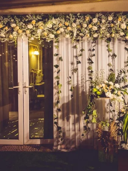 An elegant floral backdrop designed for an evening event. A cascade of white flowers and trailing ivy over sheer curtains creates a soft, romantic photo wall, complemented by modern floral stands.