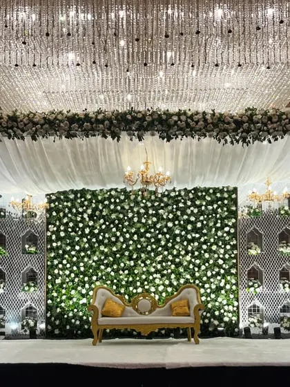 An absolutely stunning stage setup at Crowne Plaza, Okhla. A massive crystal chandelier hangs above, complementing the white floral wall, intricate side panels, and elegant white drapes.