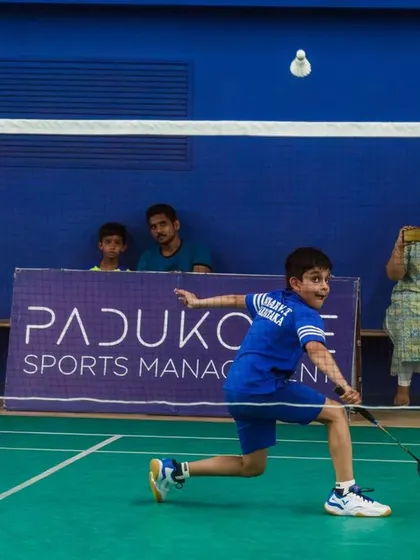 A young player lunges forward for a net shot, with parents watching in the background. We create a family-friendly environment that supports young athletes.