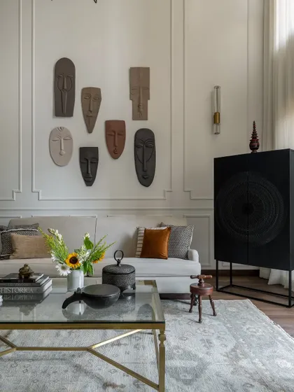 This shot highlights the curated collection of masks on the living room wall, a key feature that reflects the homeowners' travels and stories. The neutral sofa and glass table allow these personal items to stand out.