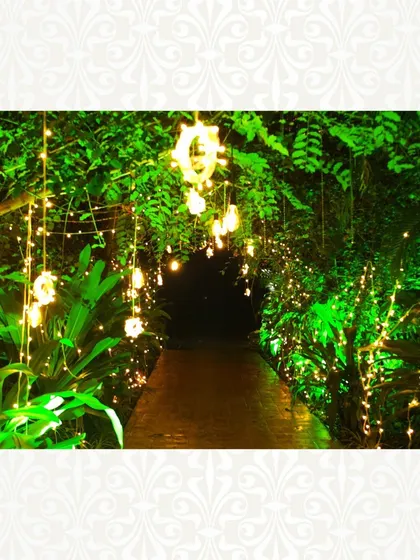 An illuminated pathway through a lush garden, with fairy lights and hanging floral arrangements creating a whimsical and dreamy atmosphere.