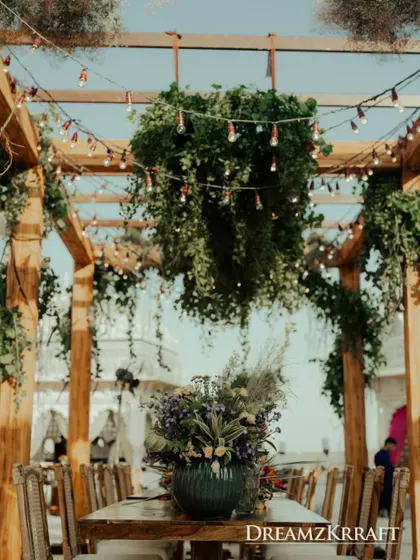 A rustic-chic dining setup under a wooden pergola draped with greenery and fairy lights, perfect for a relaxed pre-wedding meal.