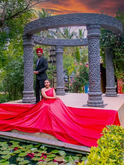A grand, wide shot of the couple at the stone structure. The red gown is spread out across the platform, creating a dramatic contrast with the ancient-style architecture and the sunset sky.