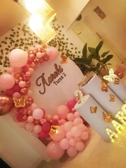 A close-up of a butterfly-themed birthday decor. This image highlights the rose gold and pink balloon garland, illuminated name, and butterfly details on the pedestals.