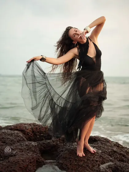 A dynamic pose with the model holding out the sheer black skirt. The movement of the fabric against the backdrop of the sea creates a beautiful and artistic fashion shot.