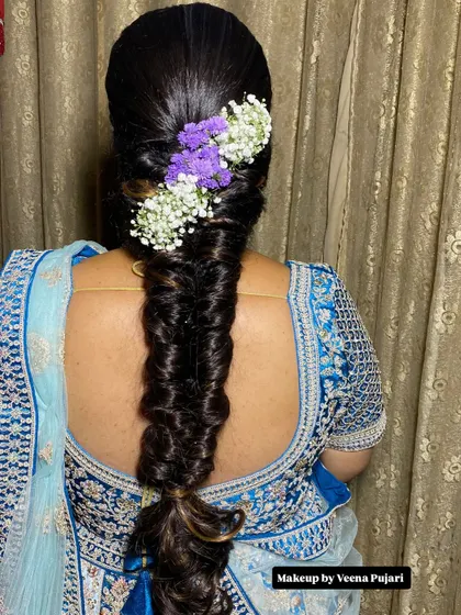 A creative and modern braided hairstyle. This fishtail braid is accented with purple and white flowers, adding a unique pop of color that complements the client's lehenga.