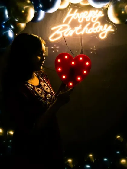 A beautiful silhouette shot of a guest holding a light-up heart prop. The 'Happy Birthday' neon sign and balloon arch create a perfect, softly lit background.