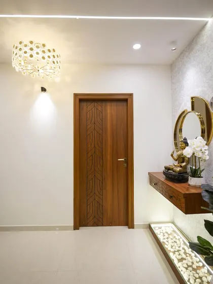 A serene foyer corner featuring a floating wooden console with under-lighting over a bed of white pebbles. The space is completed by a modern chandelier and an intricately carved wooden door, blending modern and traditional design elements.