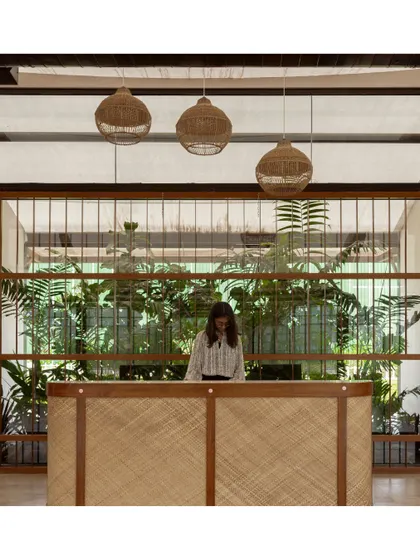 The reception desk at our Brigade Oasis project features a woven front and is set against a backdrop of lush indoor plants, creating a tropical and welcoming vibe.