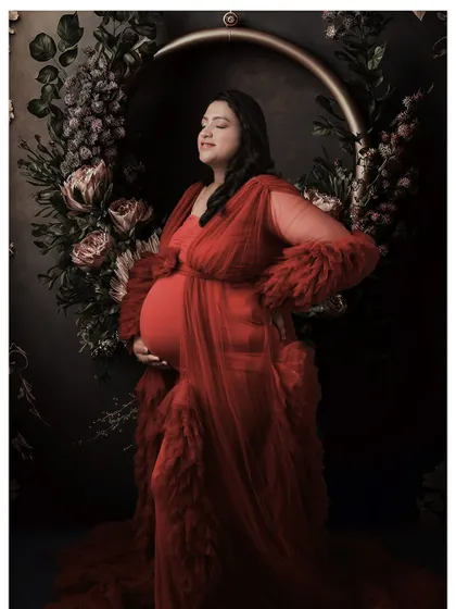 A serene portrait of a mom-to-be in a red ruffled gown, set against a dark floral backdrop. The composition is both elegant and powerful.