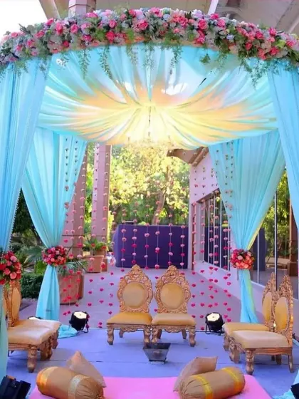 A dreamy mandap setup with pastel blue drapery and golden chairs. The canopy is adorned with pink and white flowers, and the floor is scattered with petals, perfect for a fairytale wedding.