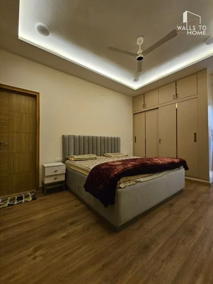A minimalist bedroom with a simple upholstered headboard, sliding wardrobes, and warm wooden flooring. The cove lighting in the false ceiling adds a soft, ambient glow.