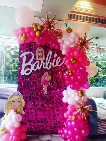 A Barbie-themed photo corner featuring a pink sequin panel, Barbie branding, and a pink and gold balloon garland.
