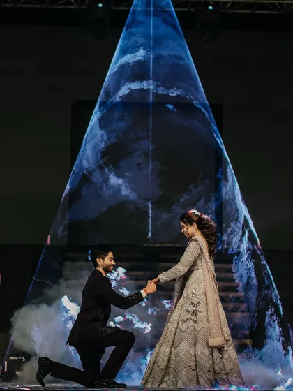 A dramatic and romantic proposal moment on the Sangeet stage. The groom kneels before his bride, framed by a stunning, futuristic light projection that creates a truly unique and memorable image.
