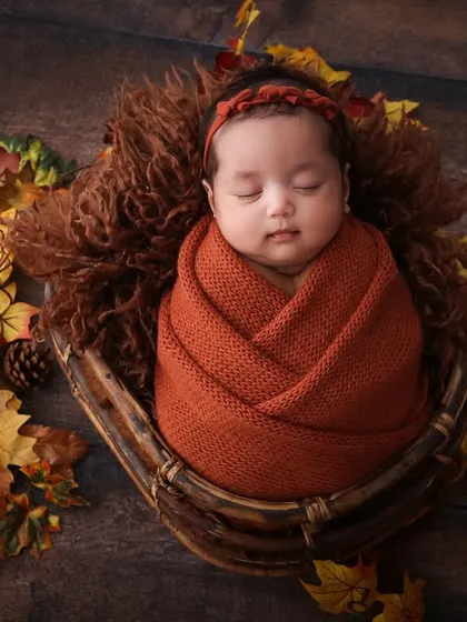 Cozy autumn vibes for a new arrival. This newborn is wrapped snugly in a rust-colored swaddle, nestled in a basket with autumn leaves. The warm colors and textures create a beautiful, seasonal portrait.