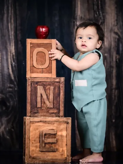 A baby boy standing next to rustic wooden "ONE" blocks, marking his first birthday.