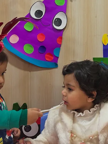 During a birthday celebration, one child gently offers a bite of cake to the birthday girl, a beautiful moment of early friendship and sharing.