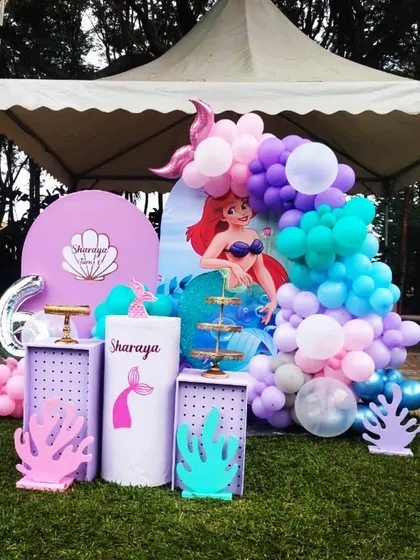 An outdoor mermaid theme for Sharaya's 6th birthday. We used a tent as the base and decorated it with an Ariel backdrop, a purple and teal balloon garland, and other under-the-sea elements.