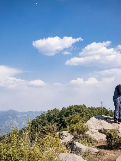 A romantic gesture on a mountaintop, with him kissing her hand against a vast blue sky filled with fluffy clouds. This wide shot captures a feeling of being on top of the world.