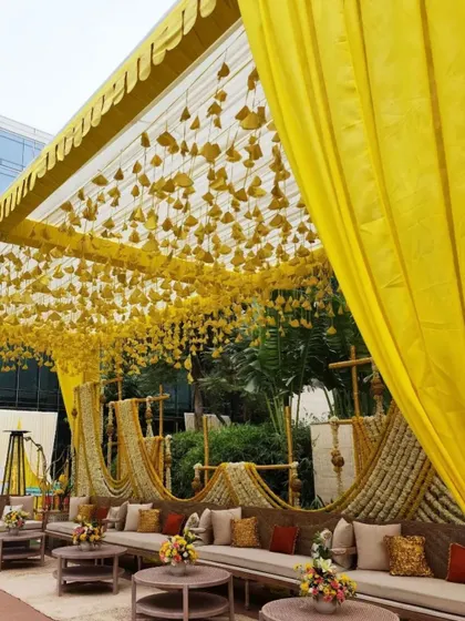A comfortable and stylish guest seating area for a Haldi ceremony. The decor uses traditional yellow drapes and hanging floral elements for a classic look.