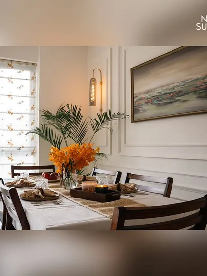 A close-up of the dining table, styled with a simple white cloth, a natural runner, and a vase of bright orange orchids. The warm lighting and classic wooden chairs create an inviting setting for any meal.