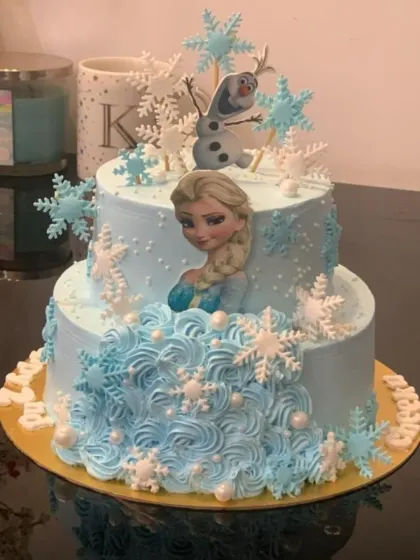 A magical two-tier Disney Frozen cake for a birthday celebration. This design features Elsa and Olaf, surrounded by a cascade of delicate, edible snowflakes and beautiful blue icing rosettes.