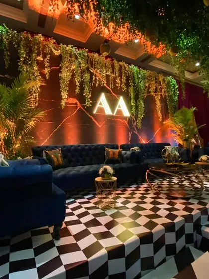 The main stage for the enchanted forest sangeet, with a checkered floor, plush blue sofas, and a backdrop of hanging vines and a neon monogram.