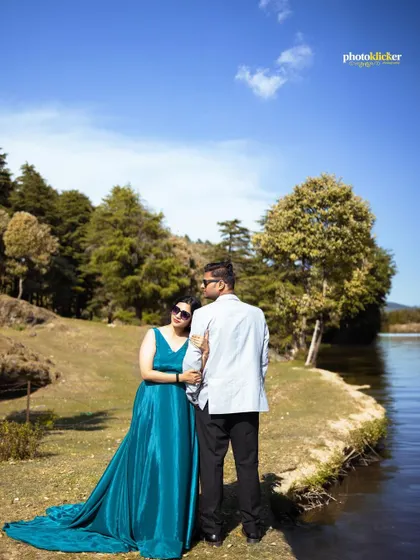 A couple enjoys a quiet moment by a mountain lake, with a clear blue sky and lush green trees creating a picturesque backdrop.