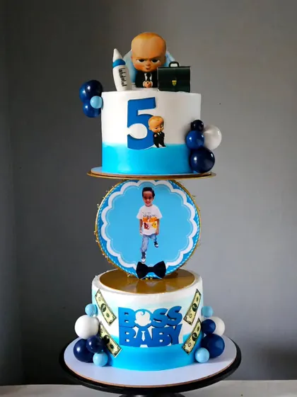 A unique "hoop" cake design for a Boss Baby theme. This modern setup features two separate tiers connected by a decorative hoop, with a photo of the birthday boy and edible Boss Baby elements.