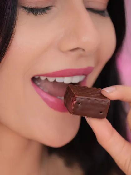 A close-up lifestyle shot of a model about to enjoy a piece of chocolate. The focus on the lips and the product creates a sensual and indulgent feel, suitable for beauty or confectionery brands.