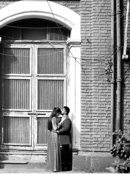 A timeless black and white pre-wedding photo of a couple sharing a kiss on the forehead against a rustic, architectural backdrop in Mumbai.