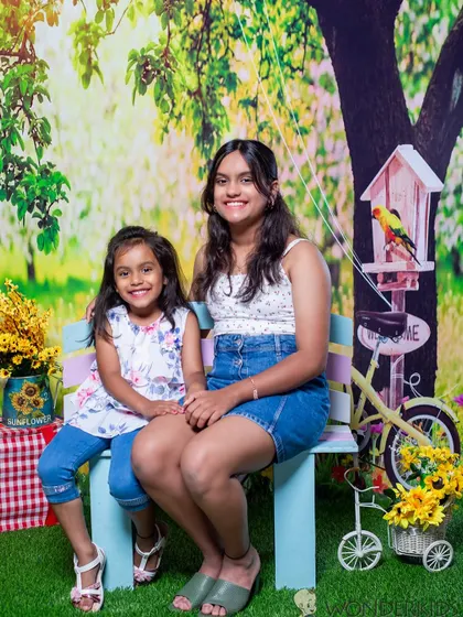 Two sisters sitting together on a bench in a garden-themed studio set. The older sister has her arm around the younger one, showing their close and protective bond.