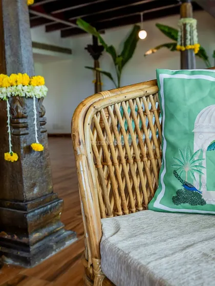 A detail of our Haldi decor, where a traditional stone pillar is adorned with marigold garlands, next to a cane sofa with a custom peacock-print cushion.
