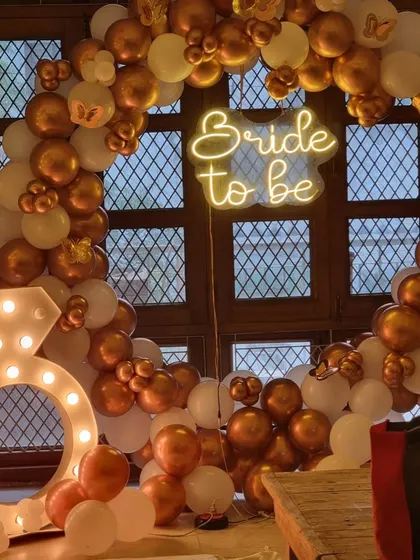 Celebrate the bride-to-be in style. This chic bachelorette party decoration features a "Bride to be" neon sign on a gold and white balloon arch, complete with a large light-up diamond ring prop for fun photos.