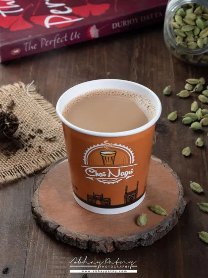 A simple cup of chai, styled to feel warm and inviting. The scattered cardamom pods and loose tea leaves hint at the authentic, spiced flavor.