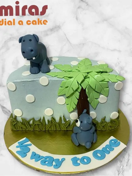 A cute "Half way to one" cake for a 6-month celebration. This rectangular cake features adorable hippo figurines and a palm tree, marking the halfway point to the big first birthday.