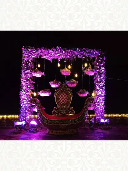 A magical baby shower setup for the mother-to-be. The decor features a royal red boat-shaped seat under a canopy of purple flowers and hanging lights, creating a dreamy and enchanting atmosphere.