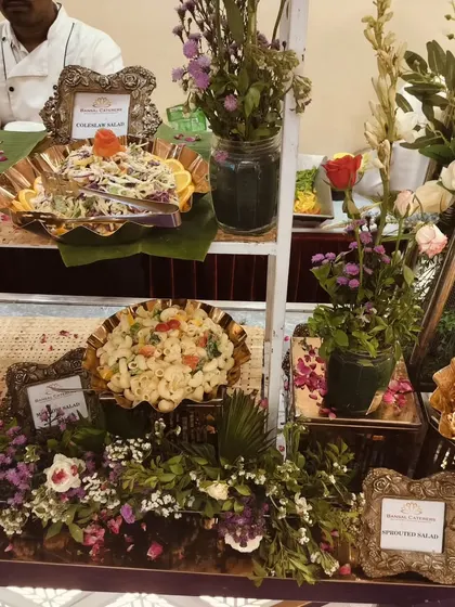 A multi-level salad display featuring coleslaw, macaroni salad, and other varieties, decorated with flowers and banana leaves.